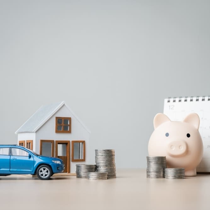 Tirelire cochon, maison, voiture bleue et piles de pièces sur un bureau pour symboliser l'épargne et le budget.